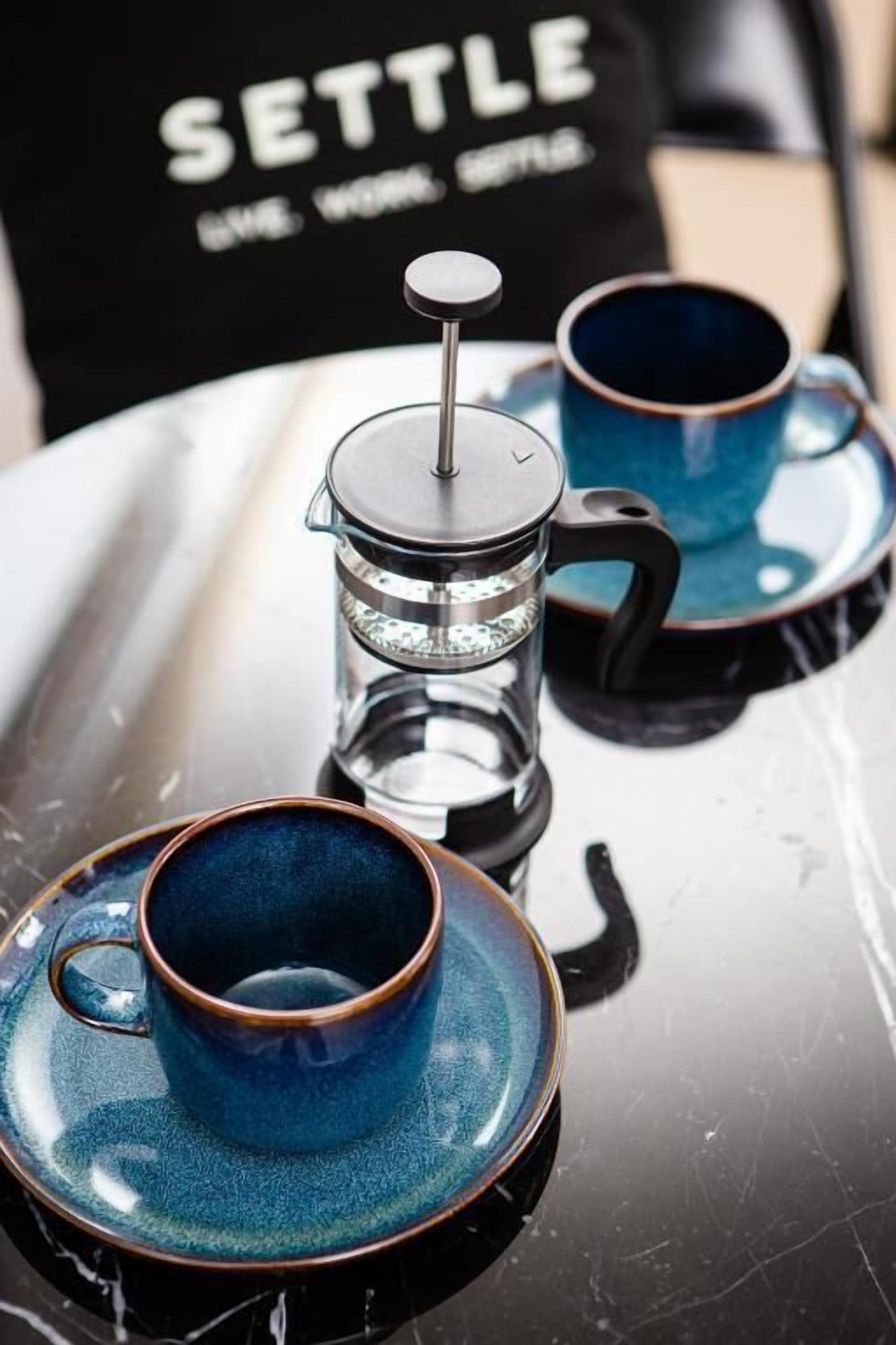 Een koffiezetapparaat met een glazen kan en twee blauwe kopjes op een reflecterend oppervlak.