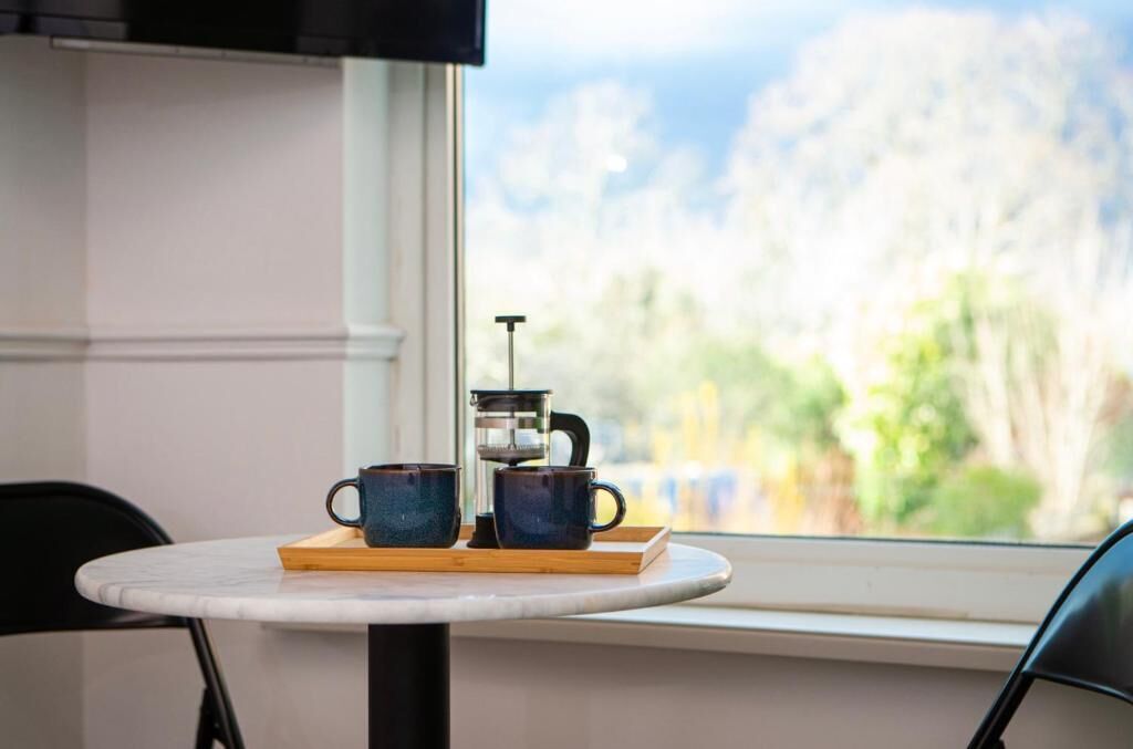 A table with two blue cups and a coffee maker by a window with a view of trees.