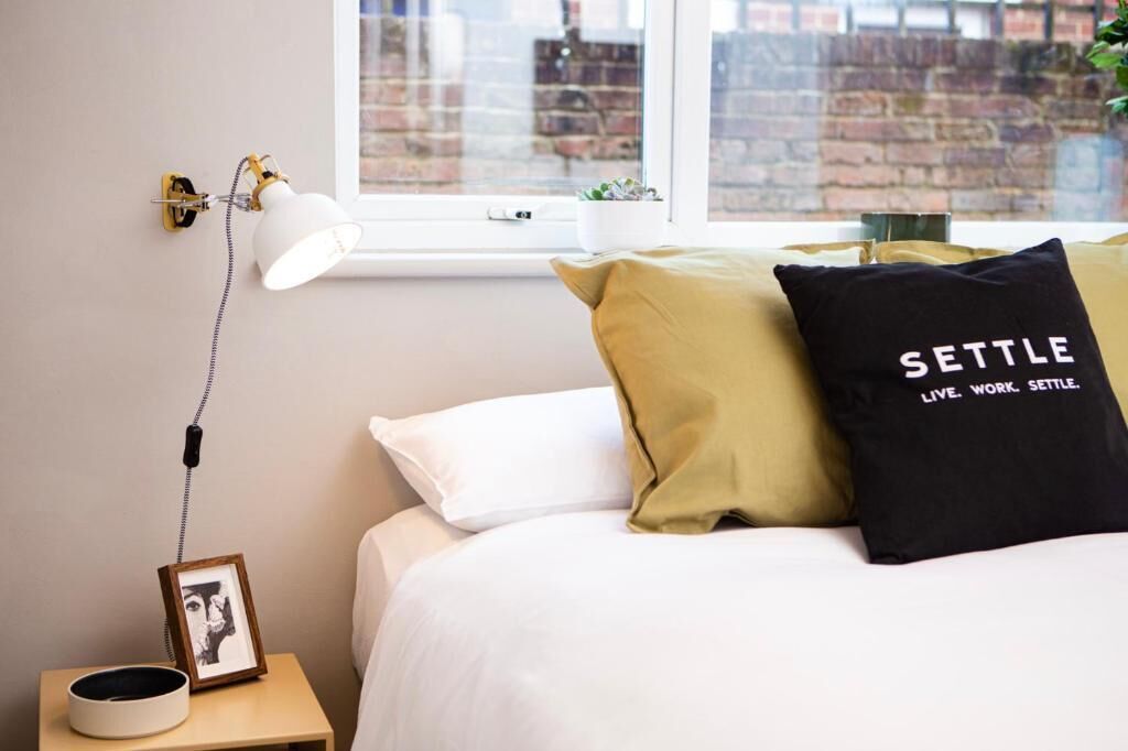 A bedroom with a bed, a bedside lamp, a framed picture, and a pillow with text.