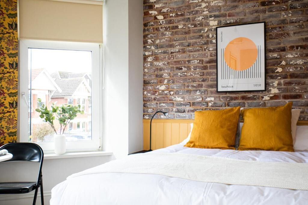 Une chambre avec un mur en briques, un lit avec une literie blanche, une fenêtre donnant sur des maisons et un tableau encadré accroché au mur.