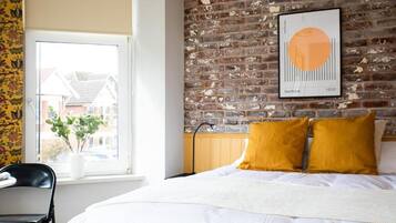 A bedroom with a brick wall, a bed with white bedding, a window with a view of houses, and a framed picture on the wall.