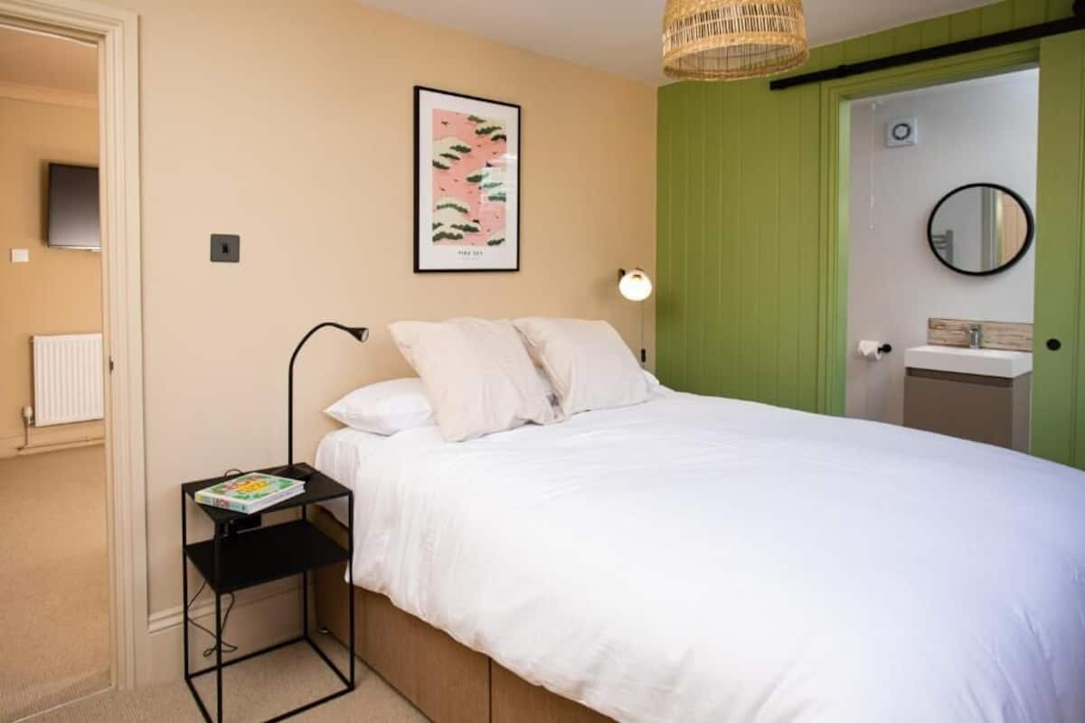 A bedroom with a bed, a bedside table with a book, a wall-mounted lamp, a framed picture, a green door, and a round mirror.