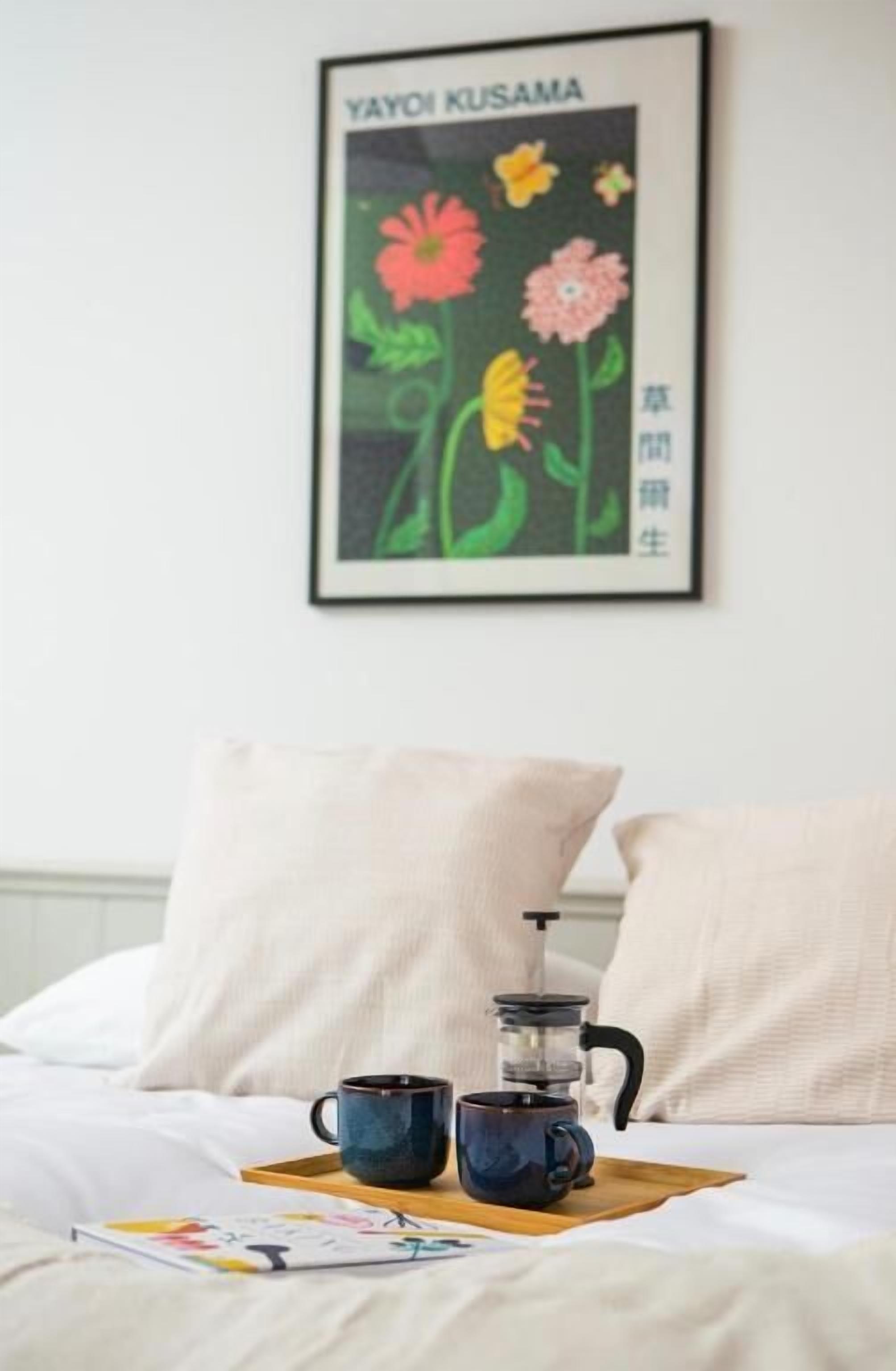 A bed with a tray holding two cups and a coffee maker, a framed picture on the wall, and a pillow.