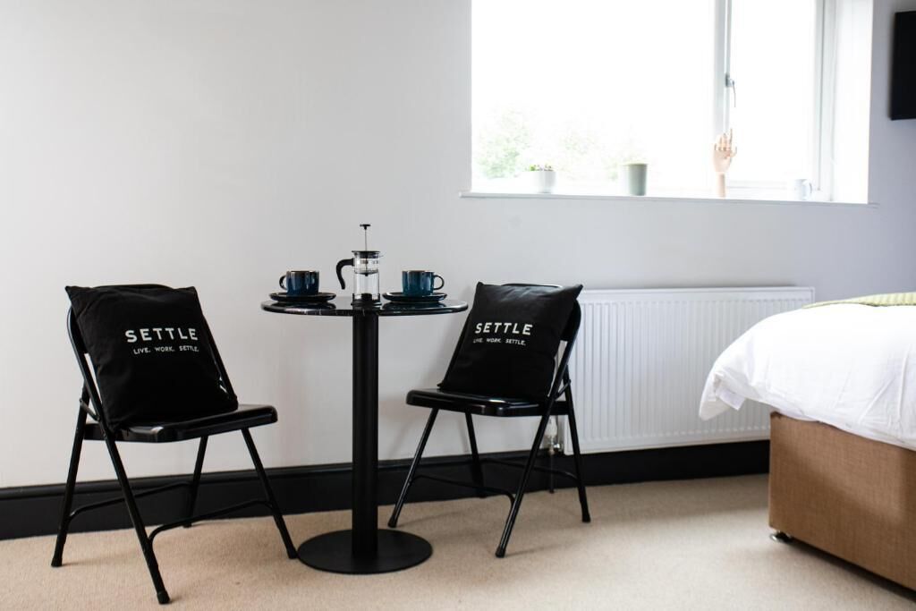 A room with a bed, two black chairs with 'SETTLE' text, a small round table with a coffee pot, cups, and a kettle.