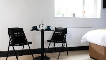 A room with a bed, two black chairs with 'SETTLE' text, a small round table with a coffee pot, cups, and a kettle.
