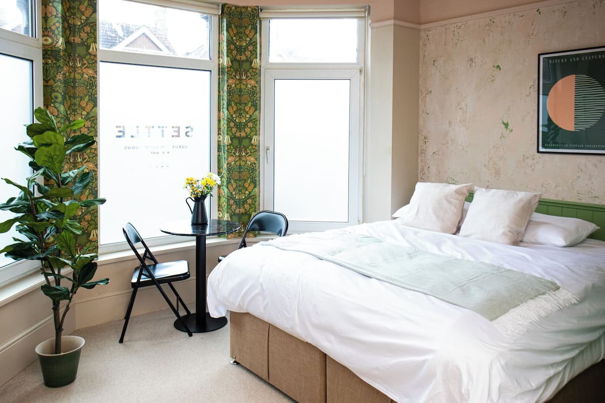 A bedroom with a bed, a chair, a table, a potted plant, and a window with curtains.