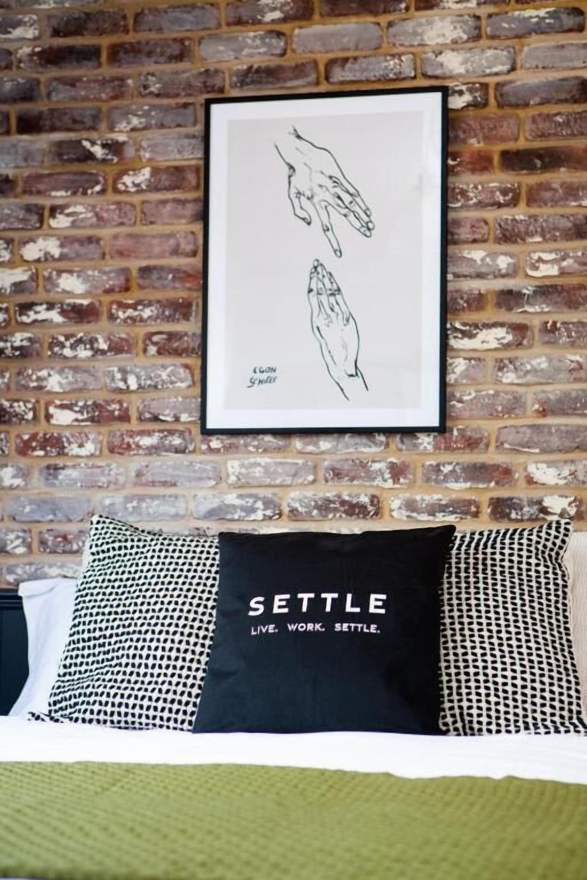 Un dibujo enmarco de manos sobre un muro de ladrillo, un cojín negro con el texto 'SETTLE LIVE. WORK. SETTLE.' y un cojín estampado sobre una cama.
