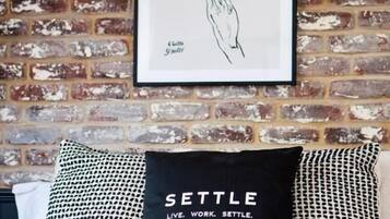 Un dibujo enmarco de manos sobre un muro de ladrillo, un cojín negro con el texto 'SETTLE LIVE. WORK. SETTLE.' y un cojín estampado sobre una cama.