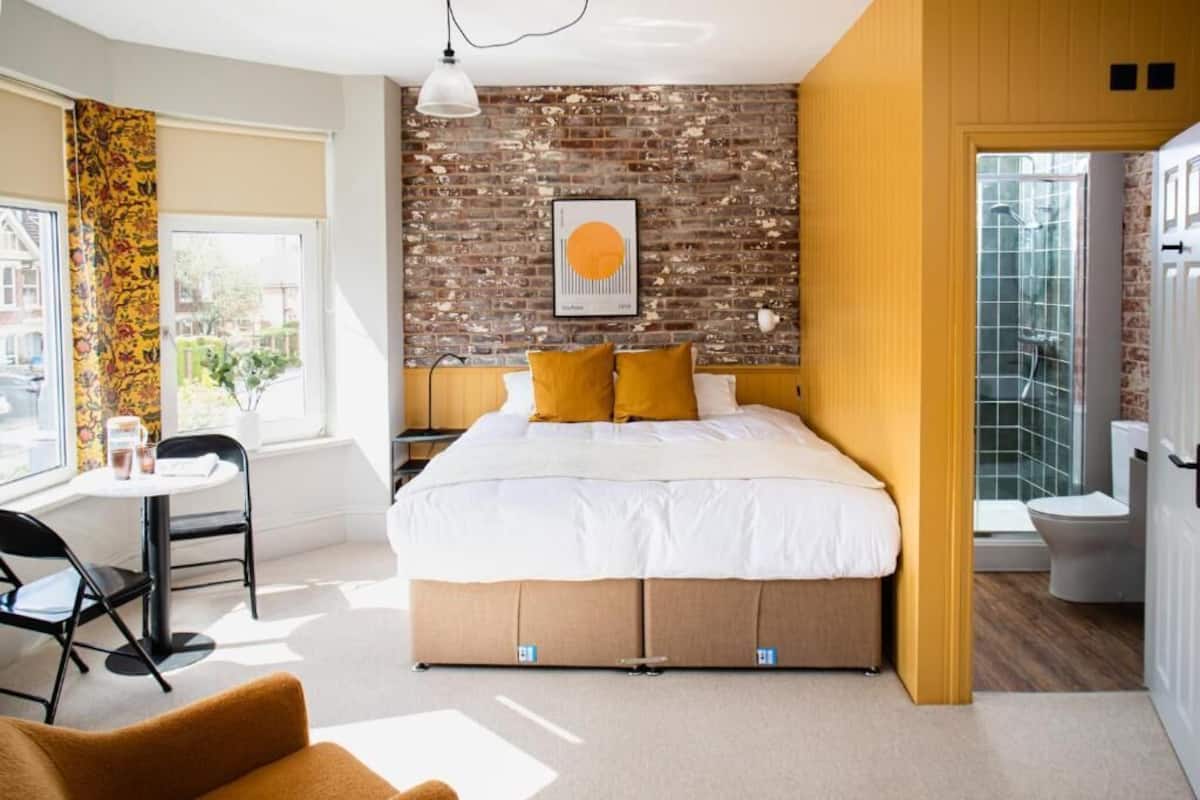 A bedroom with a brick wall, a bed with white bedding and yellow pillows, a small table with chairs, and a window with curtains.