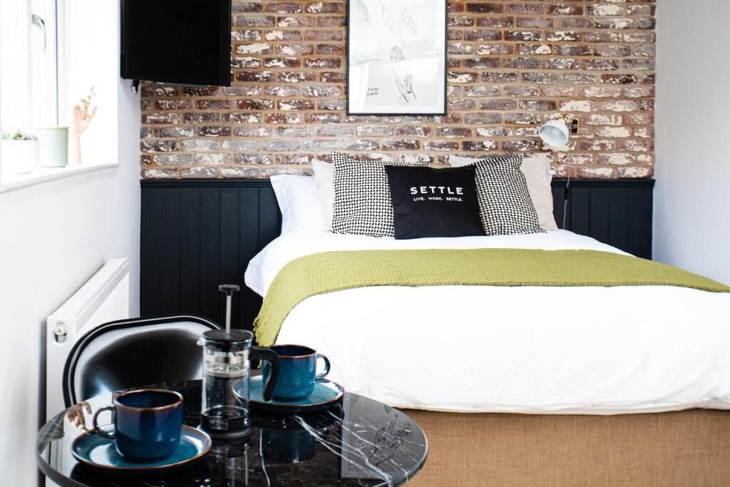 A room with a brick wall, a bed with a white and green bedspread, a black and white pillow, a black coffee table with cups and a coffee pot, and a framed picture on the wall.
