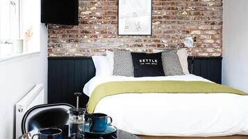 A room with a brick wall, a bed with a white and green bedspread, a black and white pillow, a black coffee table with cups and a coffee pot, and a framed picture on the wall.