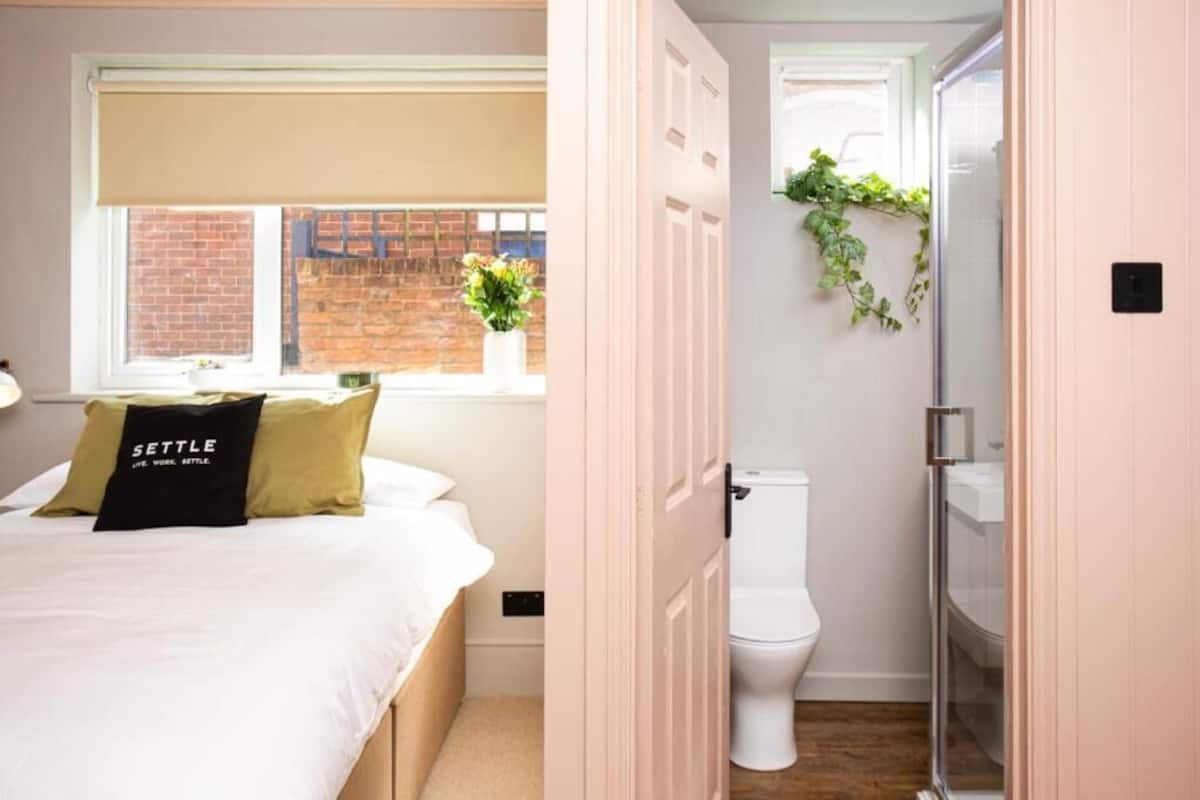 A bedroom with a bed, a window with a view of a brick wall, and a toilet in the adjacent bathroom.