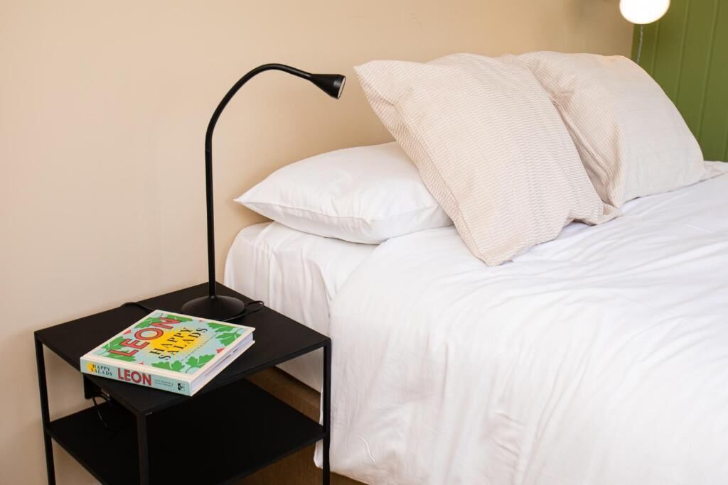 A bed with white bedding, a black nightstand with a book titled 'Leon Happy Salads,' and a black desk lamp.