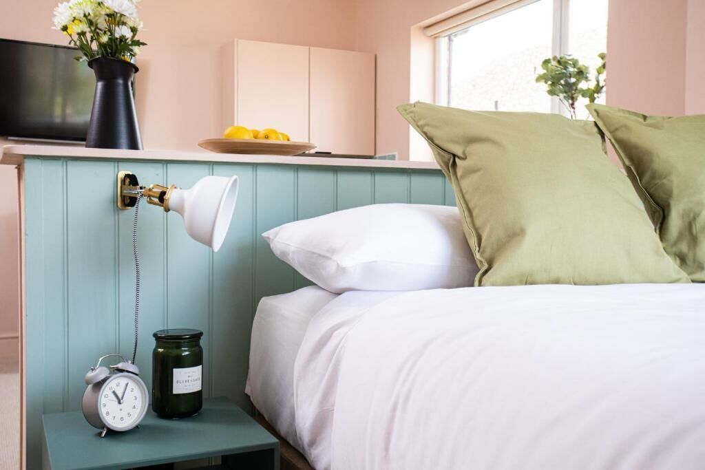 A bedroom with a bed, a nightstand with a clock and a jar, a lamp, a vase with flowers, and a window.