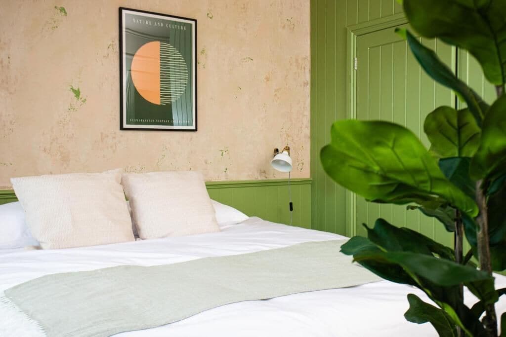 A bedroom with a bed, pillows, a framed picture, a green wall, and a plant.