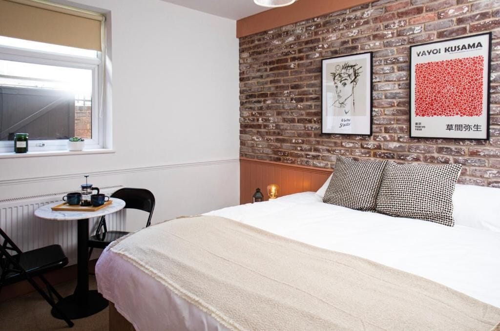 Ein Hotelzimmer mit Backsteinwand, einem Bett mit gemusterten Kissen, einem kleinen Tisch mit Teekanne und gerahmten Kunstwerken.