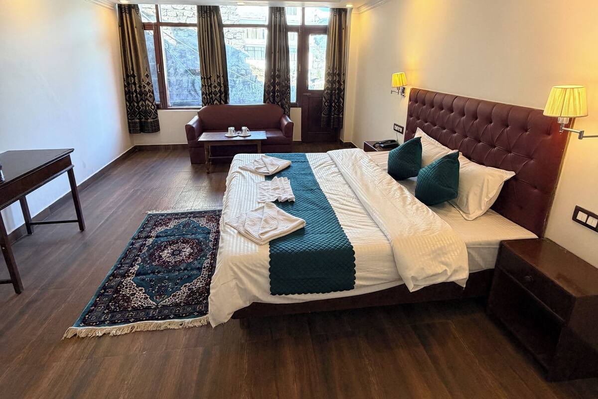 A hotel room with a large bed, a sitting area with a sofa and table, and a view of the outside through large windows.