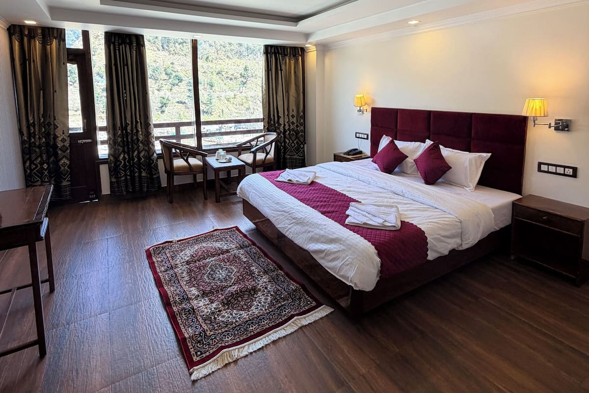 A hotel room with a large bed, a desk, a chair, a small table, a nightstand, and a window with curtains.