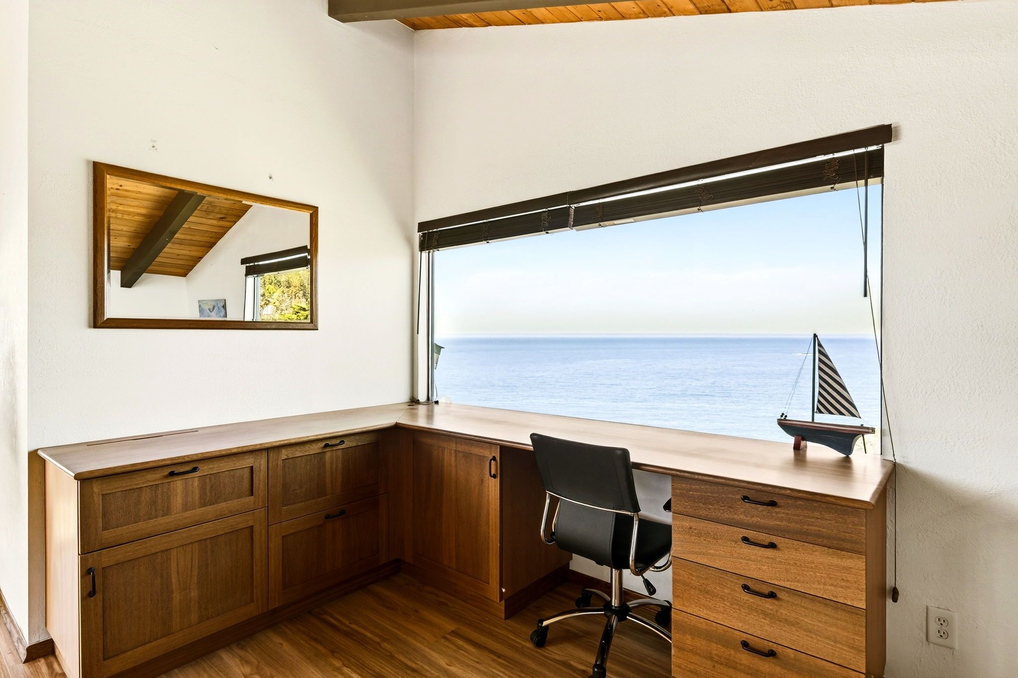 A corner desk with a view of the sea, a small sailboat model, and a mirror on the wall.