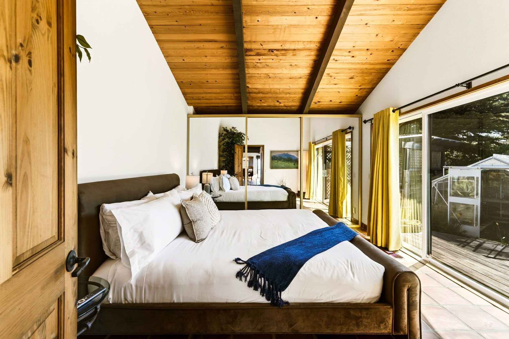 A bedroom with a wooden ceiling, a large bed with white bedding, a blue throw blanket, and a view of the outdoors through a sliding glass door.