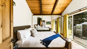 A bedroom with a wooden ceiling, a large bed with white bedding, a blue throw blanket, and a view of the outdoors through a sliding glass door.