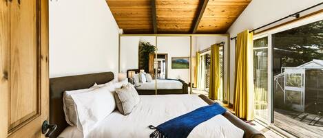 A bedroom with a wooden ceiling, a large bed with white bedding, a blue throw blanket, and a view of the outdoors through a sliding glass door.