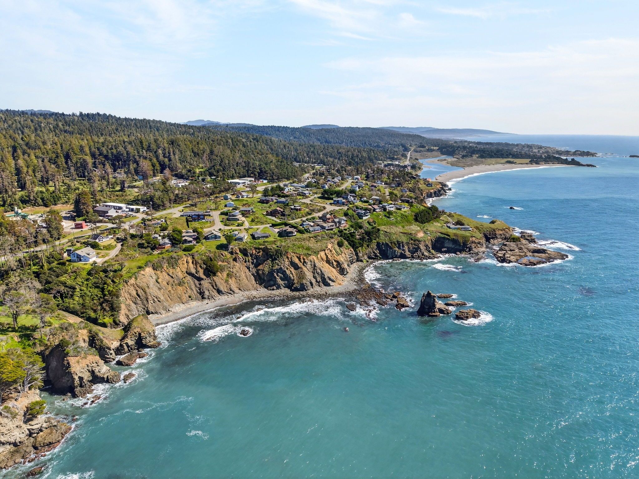 A coastal residential area with houses along the shoreline, a rocky coastline, and a forested hill in the background.