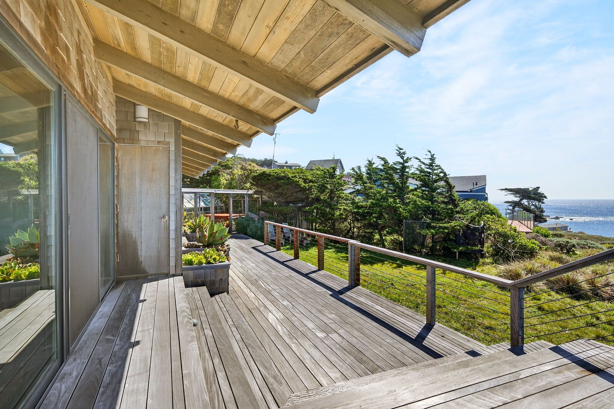A wooden deck with a railing, overlooking a lush garden and a coastal view.