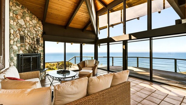 Amplia habitación con pared de piedra, chimenea, muebles de mimbre y grandes ventanas con vistas al mar.