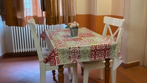 Un comedor con una mesa cubierta por un mantel estampado, dos sillas blancas y un florero con flores.