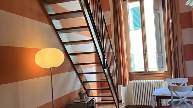 Una sala de estar con sofá, lámpara, mesa de comedor con sillas, escalera y ventana con cortinas.