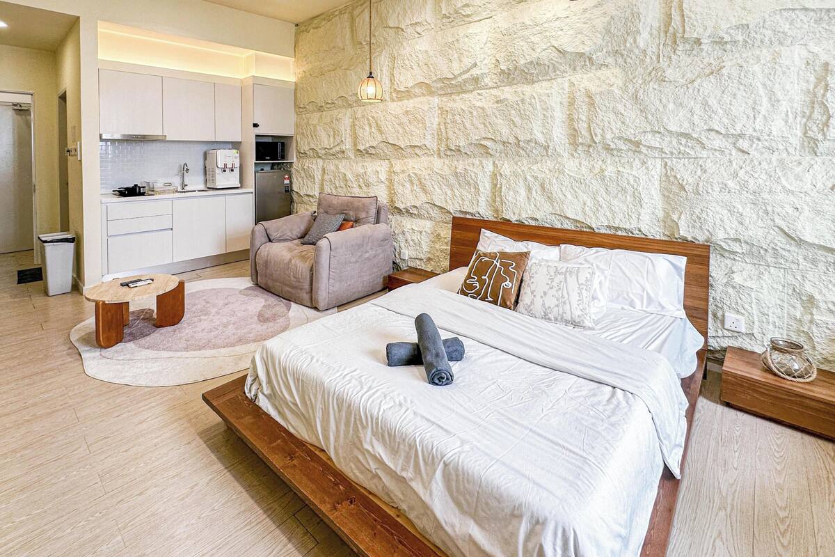 A bedroom with a large bed, a small seating area, a kitchenette, and a stone-textured wall.