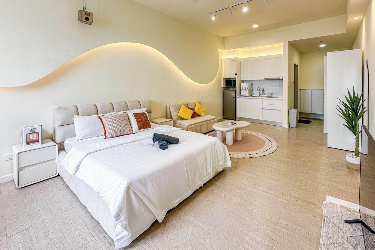 A modern hotel room with a large bed, a sofa, a small table, a kitchenette, and a TV.
