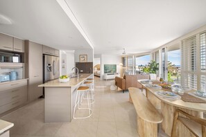 A modern kitchen with a central island, bar stools, and a dining area with a view. - Salt & Sky - Stylish Apartment with Pool & Spa (Caloundra Region)
