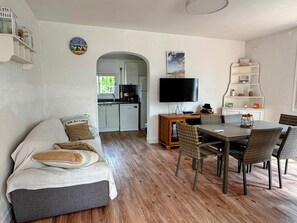 Ein modernes Wohnzimmer mit einer Couch, einem Esstisch, Stühlen, einer Küche im Hintergrund und einem an der Wand befestigten Fernseher.