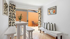 Um quarto com um banco branco, uma mesa pequena, um sofá de vime com almofadas, uma planta em vaso e um quadro na parede.
