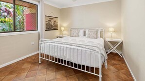 Quarto com cama de metal branca, ventilador de teto, janela com vista para vegetação e mesa de cabeceira com abajur.