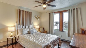 A bedroom with a bed, bedside lamps, a ceiling fan, a window with curtains, and a wooden nightstand. - Ohio Valley Home Near Franciscan University! (Steubenville)