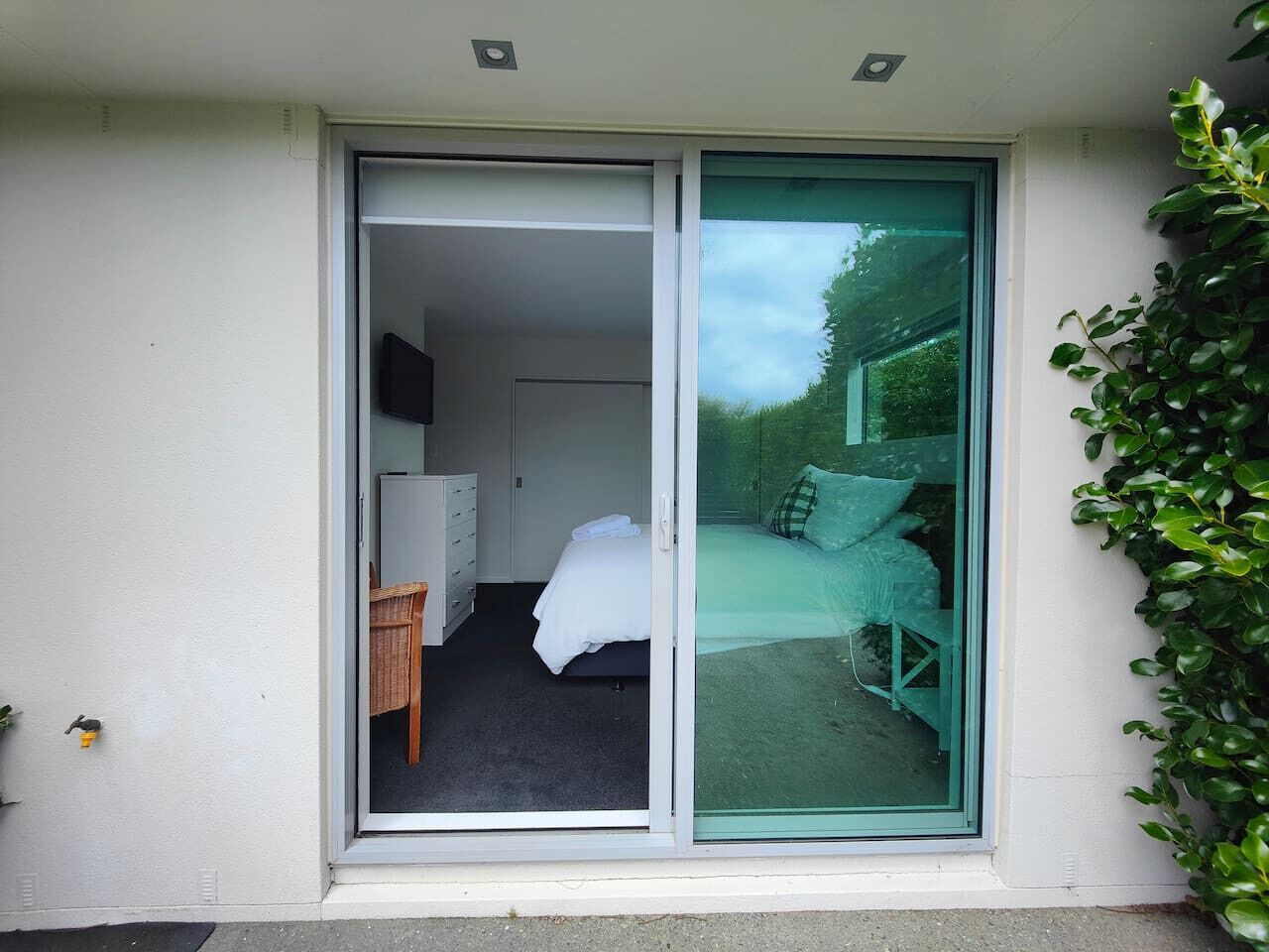 Quarto com cama, escrivaninha e cadeira, porta de correr de vidro que dá para área externa com banco e vegetação.