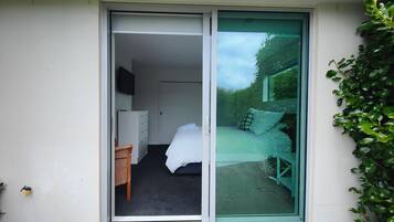 Quarto com cama, escrivaninha e cadeira, porta de correr de vidro que dá para área externa com banco e vegetação.