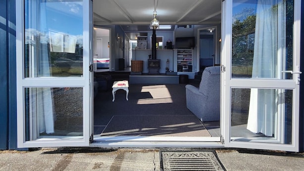 A house with a sliding glass door leading to a living room with a sofa, a small table, and a fireplace. - Blue Bach in the Loop (Te Anau)