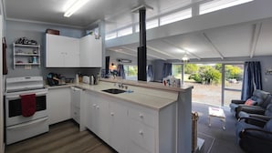 A modern kitchen with white cabinets, a stove, and a sink. There is a dining area with a table and chairs, and a view of the outdoors through large windows. - Blue Bach in the Loop (Te Anau)