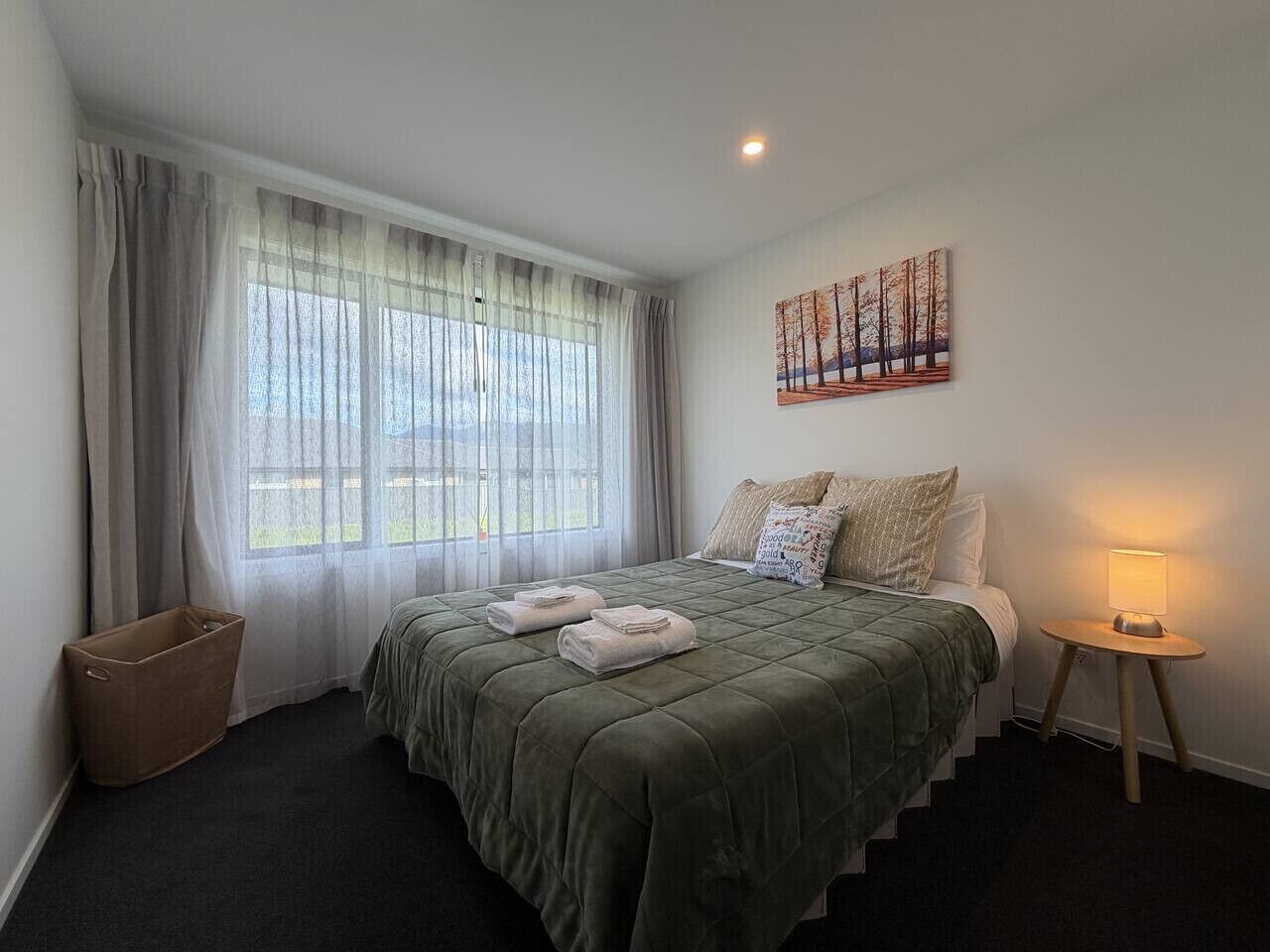 A bedroom with a bed, a nightstand, a window with curtains, a painting on the wall, and a trash bin.