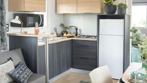 A compact kitchenette with a microwave, stove, and cabinets, a gray sofa with patterned cushions, and a dining area with a table and chairs. - Mobile home Keiheuvel (6p) (Balen)