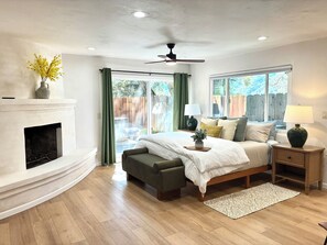 A bedroom with a fireplace, a bed with pillows and a bedside table, a window with curtains, and a ceiling fan. - Charming 1-bedroom lodge in peaceful Carmel Valley w/ Hot Tub and backyard (Carmel Valley)