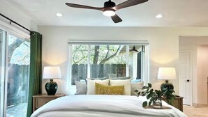 Un dormitorio con cama, dos lámparas de noche, ventilador de techo y ventana con vistas a árboles.