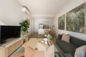 A modern living room with a flat-screen TV, a large plant, a dining area with a table and chairs, and a kitchen in the background.