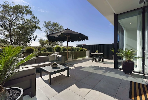 A patio with a sofa, table, and umbrella, surrounded by potted plants and a view of the sky.
