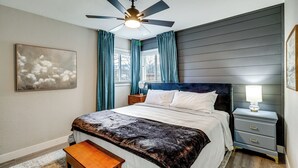 Ein Schlafzimmer mit einem Bett, einem Nachttisch mit Lampe, einem Deckenventilator und einem Fenster mit Vorhängen.