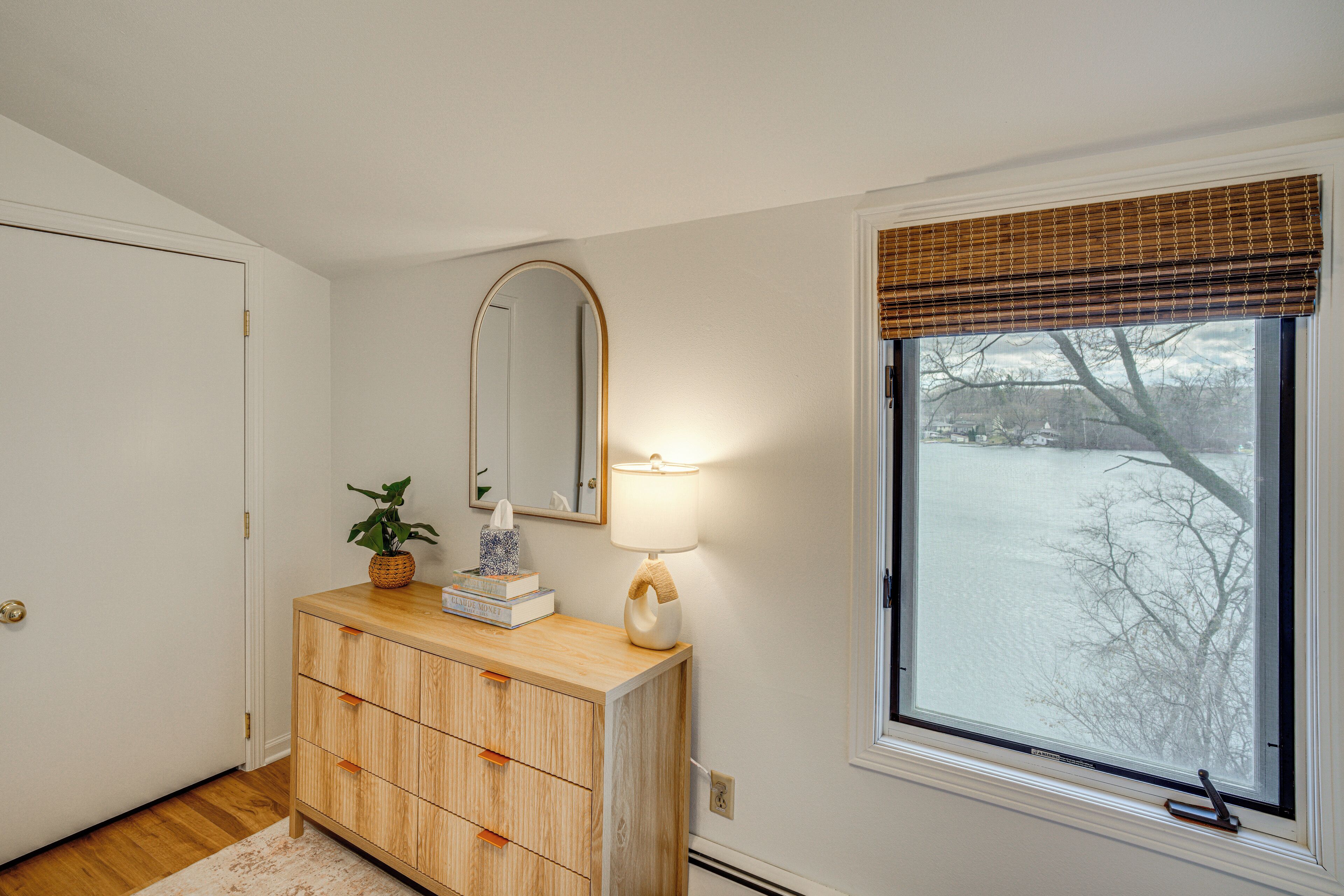 A room with a wooden dresser, a mirror, a lamp, and a window with a view of a snowy landscape.
